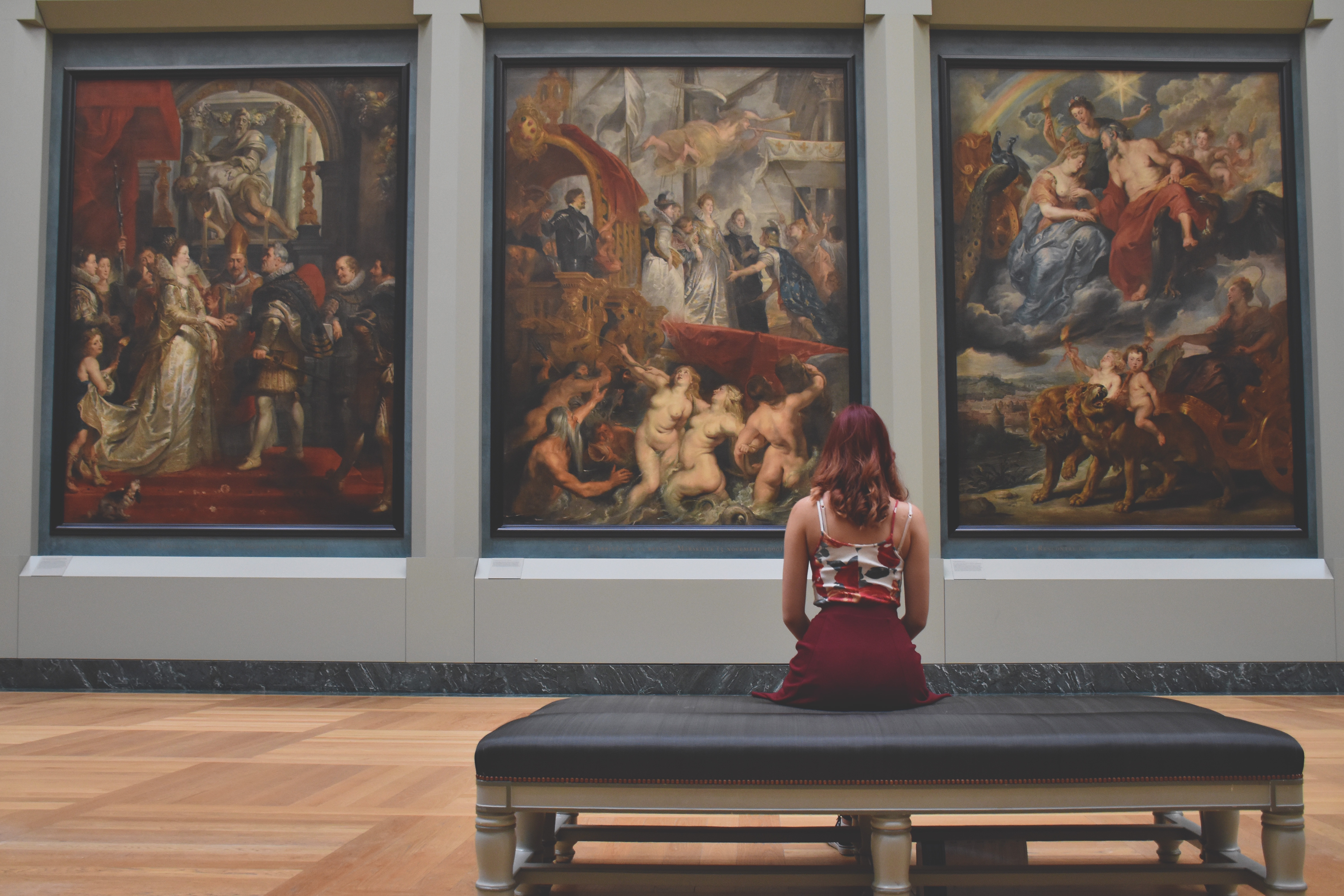 An image of a woman sitting on a bench while looking at three paintings